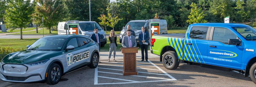 A Judd Herzer speaks at a lectern in a parking lot during a press event, surrounded by colleagues and four electric vehicles. On the left is a silver and green police SUV, and on the right is a blue and green Consumers Energy pickup truck. Two white electric vans are parked in the background.