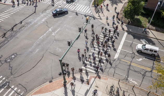 Aerial view of a busy intersection with multiple crosswalks, where a large group of pedestrians is crossing the street under sunny daylight. Several cars, bicycles, and traffic lights are visible, along with shadows cast by the people and surrounding objects.