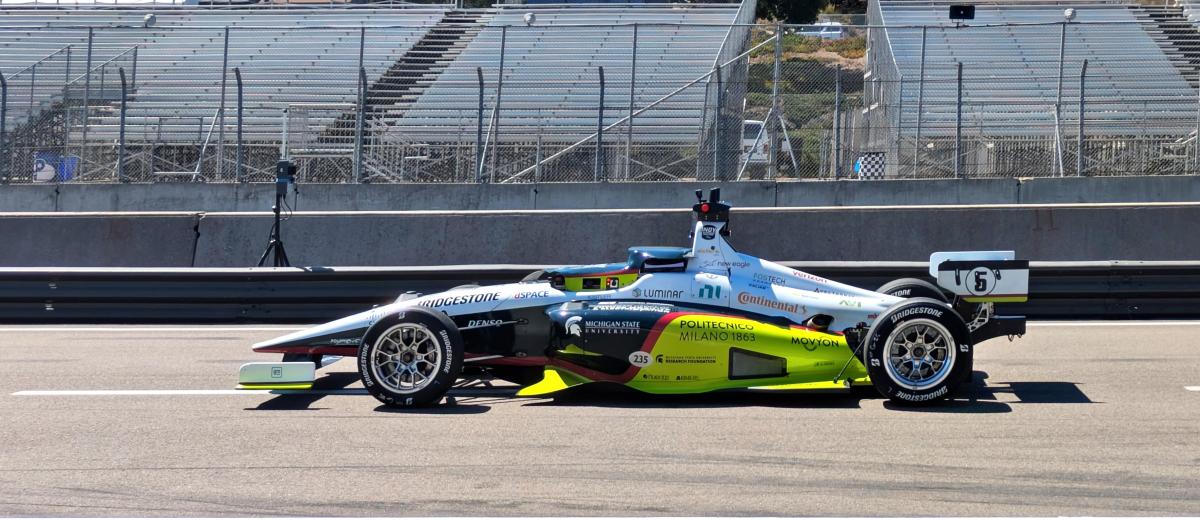 Brightly colored self-driving race car on a racetrack in front of empty grandstands.