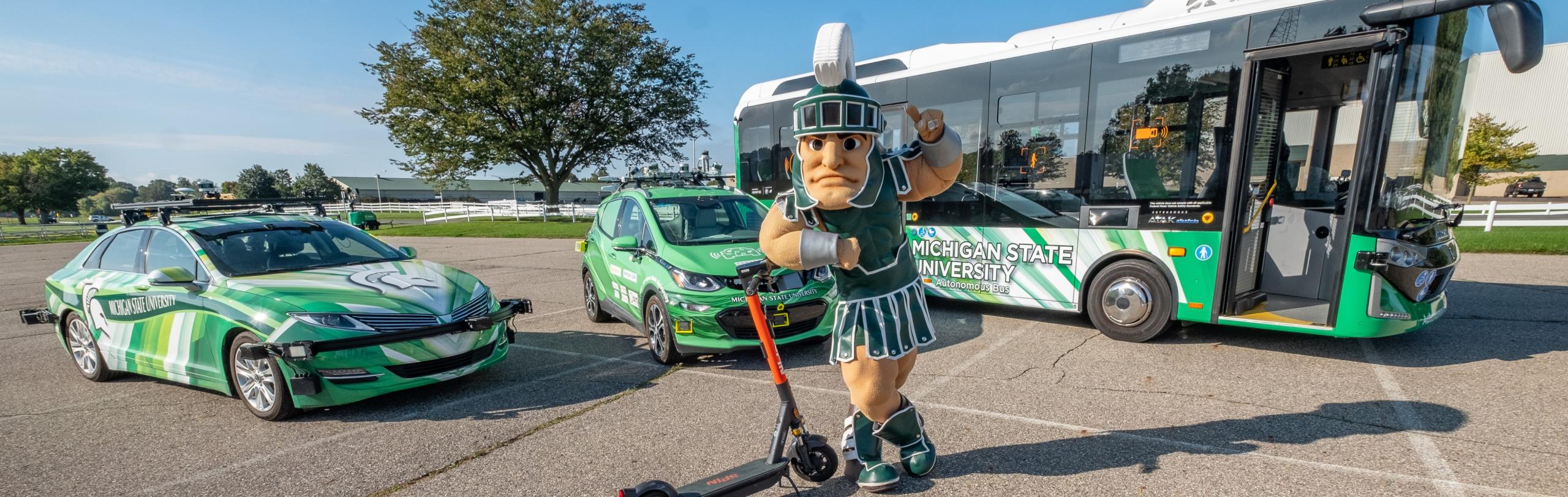 Michigan State University mascot Sparty poses with an electric scooter in front of a lineup of autonomous vehicles, including a green MSU-branded car, a compact electric vehicle, and a green-and-white autonomous bus, all parked outdoors on a sunny day.
