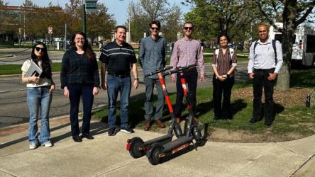 Group of seven people standing on a sidewalk near two parked Spin electric scooters on a sunny day.