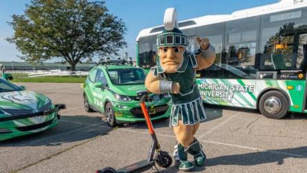 Sparty standing with a scooter in front of two autonomous cars and an autonomous bus.